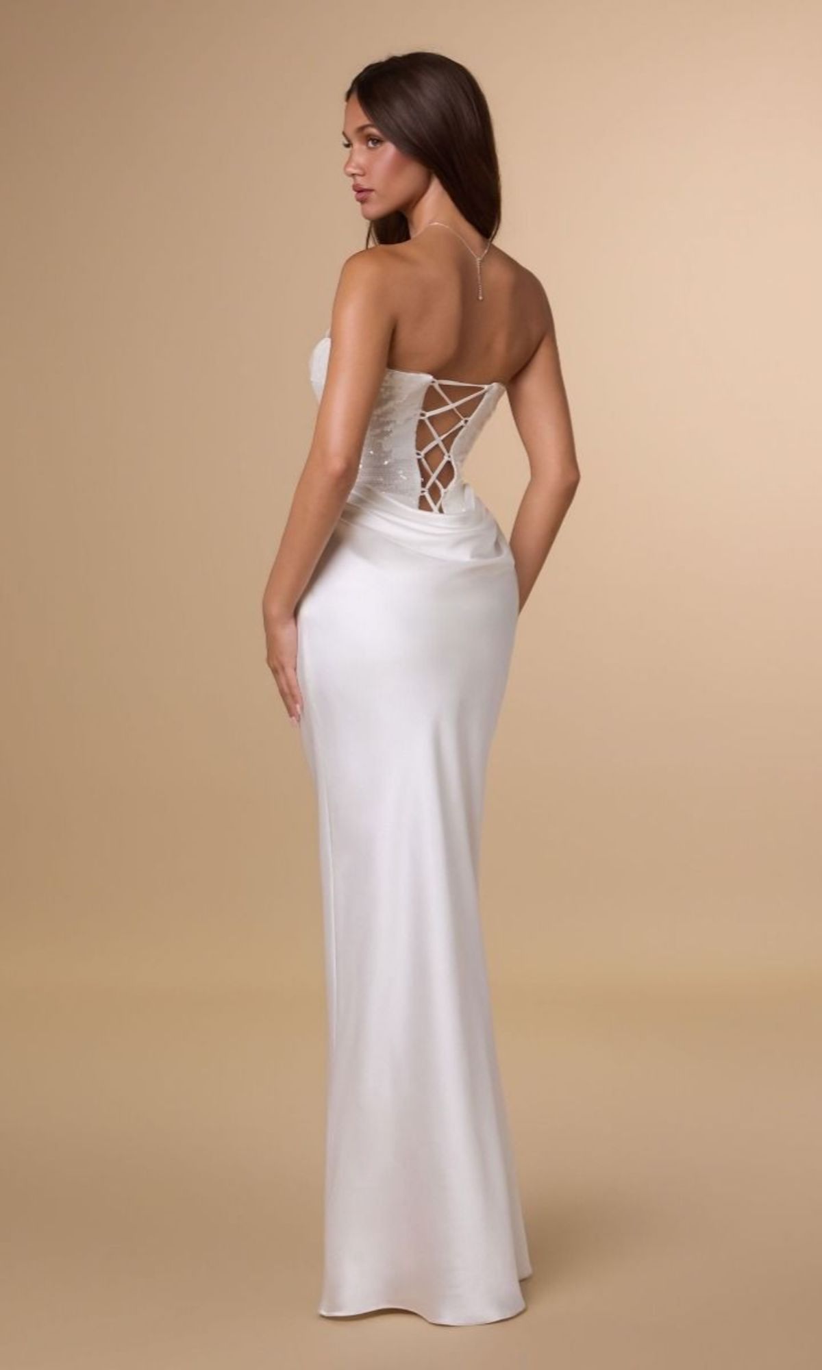 A woman wearing the Long Prom Dress: Amoris T744W, which features a white strapless design with a sequin bodice, lace-up back, and a flowing satin skirt, stands against a plain beige background facing away from the camera.